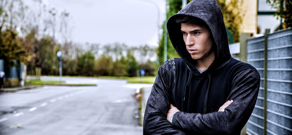 Man stood outside with a black hoodie on with his hood up and arms crossed.